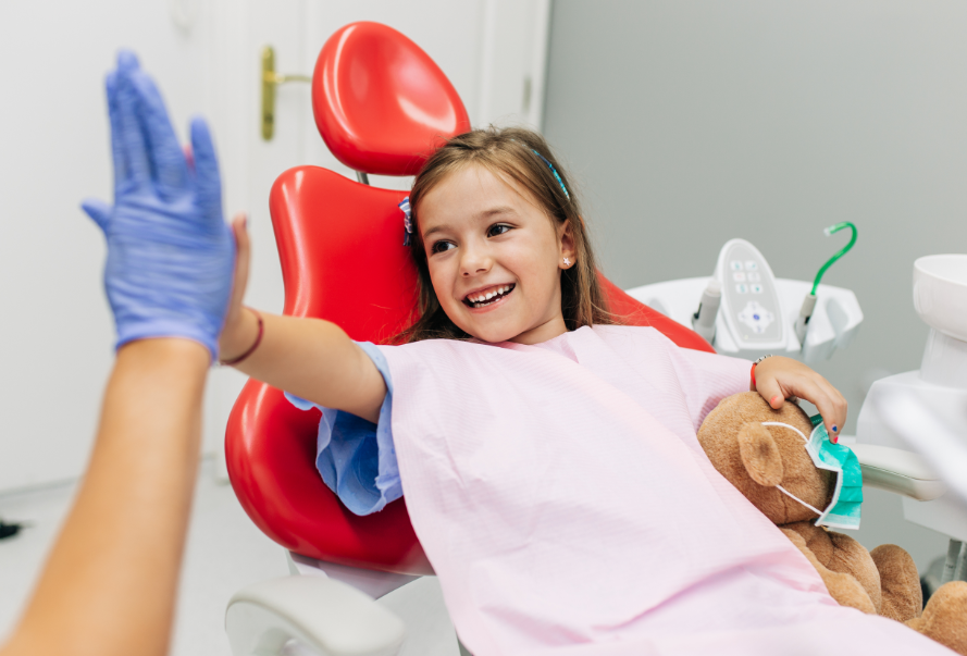 Little girl in dentist chair with teddy bear, high fiving dentist